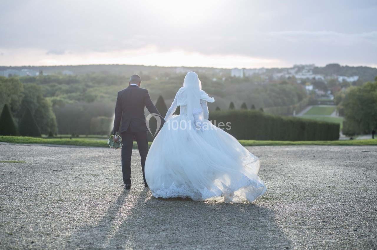 Un couple en tenue de mariage marche main dans la main sur une allée, avec un paysage verdoyant en arrière-plan.