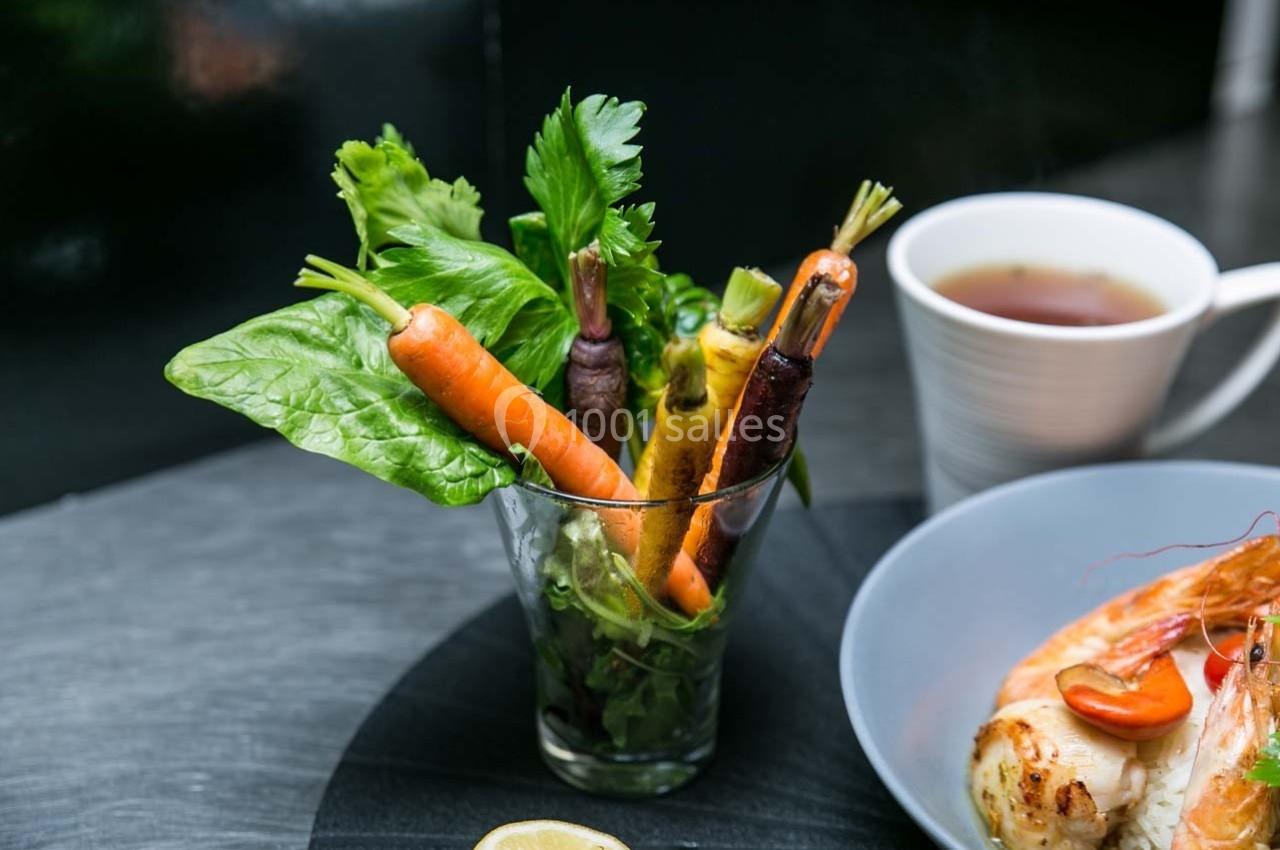 Légumes frais, dont carottes colorées et feuilles vertes, disposés dans un verre sur une table avec des plats à côté.