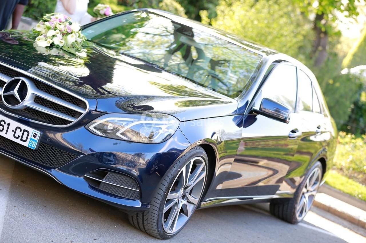 Voiture de luxe noire décorée de fleurs blanches, garée sur une route bordée de verdure.