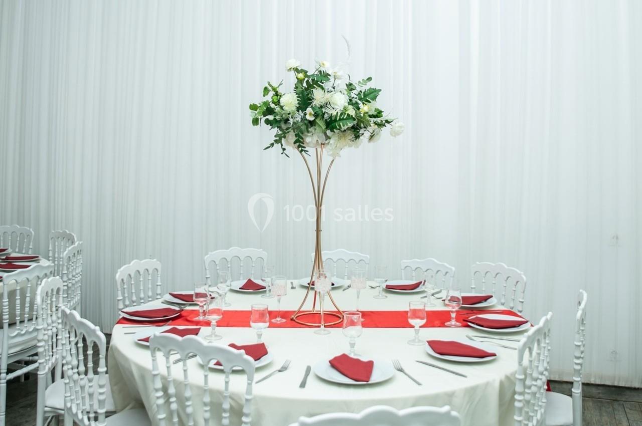 Table ronde décorée avec une nappe blanche, des serviettes rouges et un centre de table floral sur pied doré.