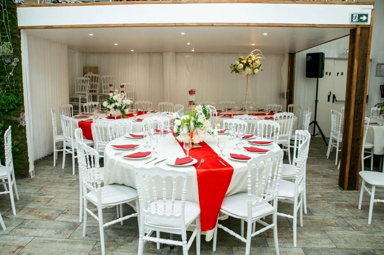 Salle de réception décorée avec des tables rondes, nappes blanches, chemins de table rouges et chaises blanches.