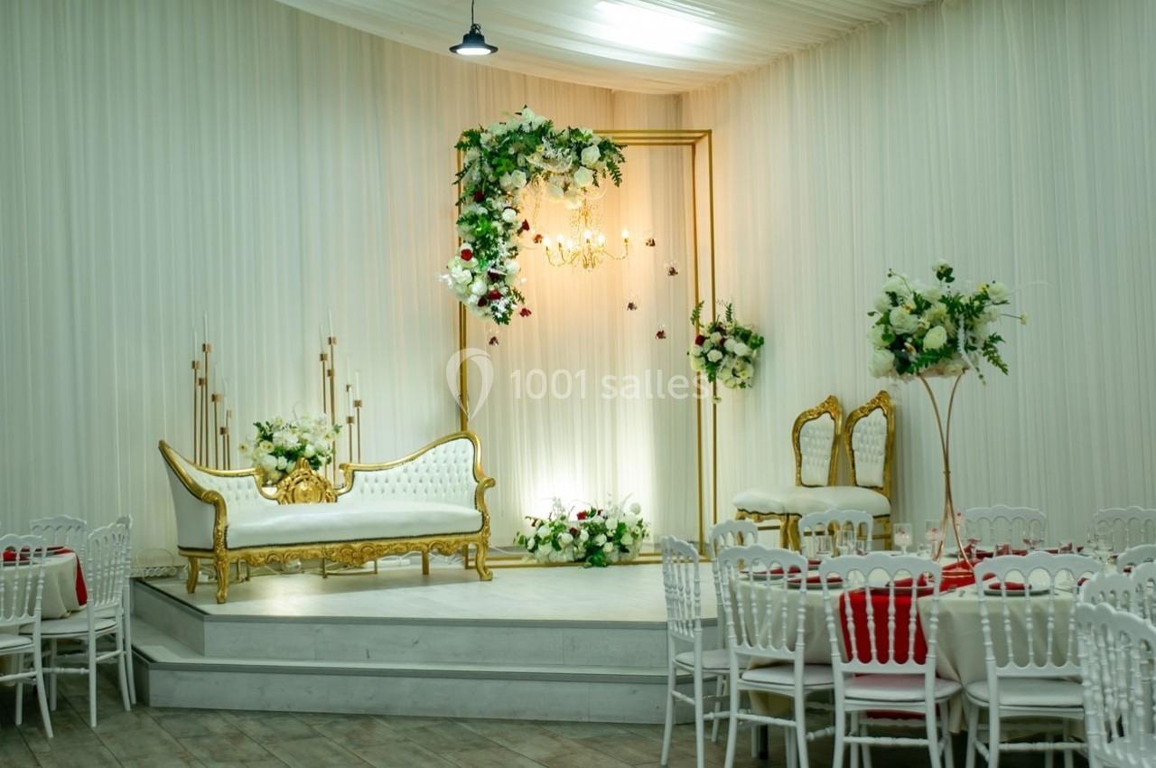 Décor de salle avec canapé doré, arche fleurie, chaises blanches et tables dressées pour un événement.