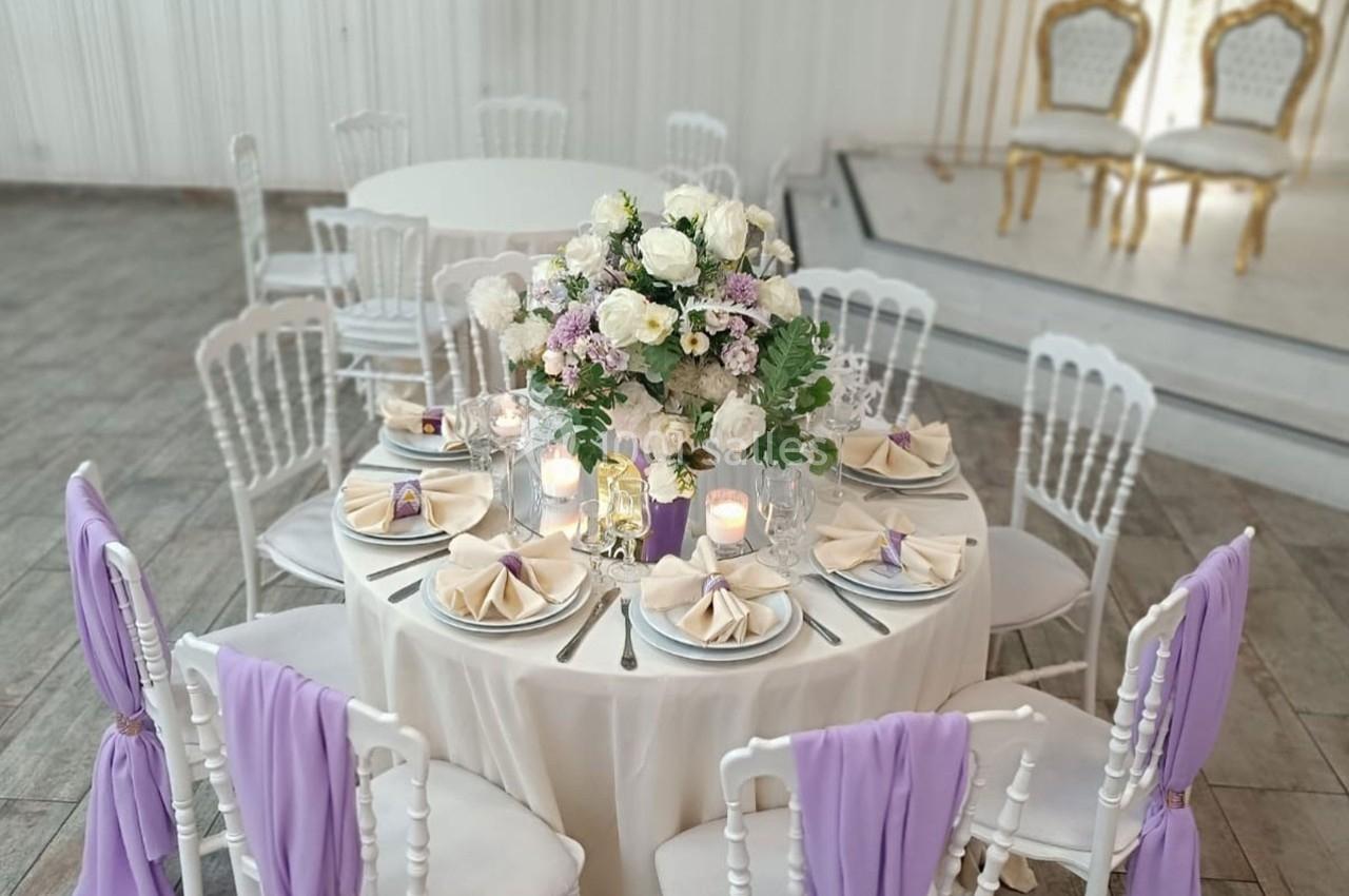 Table élégamment dressée avec des fleurs blanches et lilas, serviettes pliées et chaises décorées de tissu violet.
