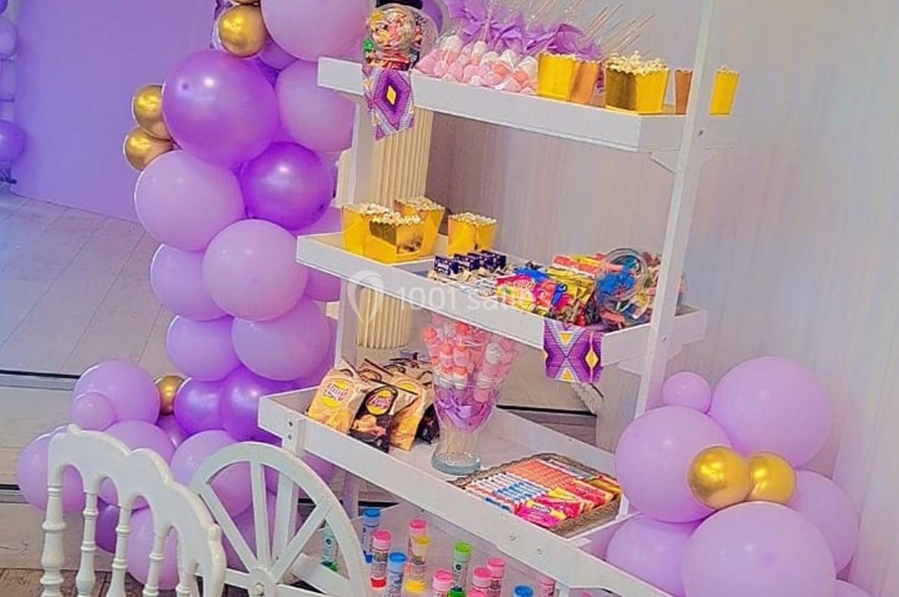 Chariot décoré de ballons violets et dorés, rempli de friandises et snacks colorés pour une fête ou célébration.