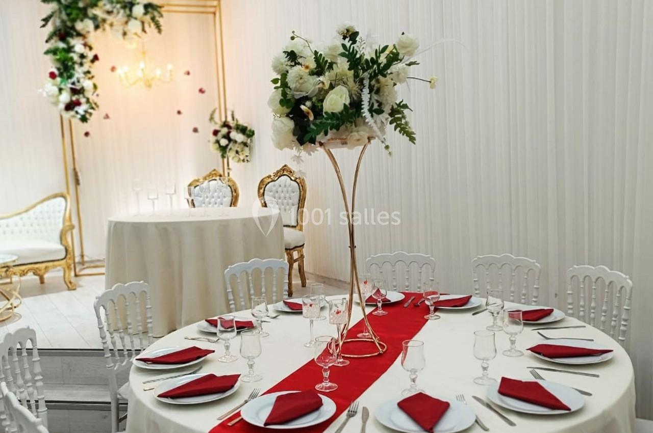 Salle de réception décorée avec des tables rondes, nappes blanches, chemins de table rouges et un centre floral élégant.