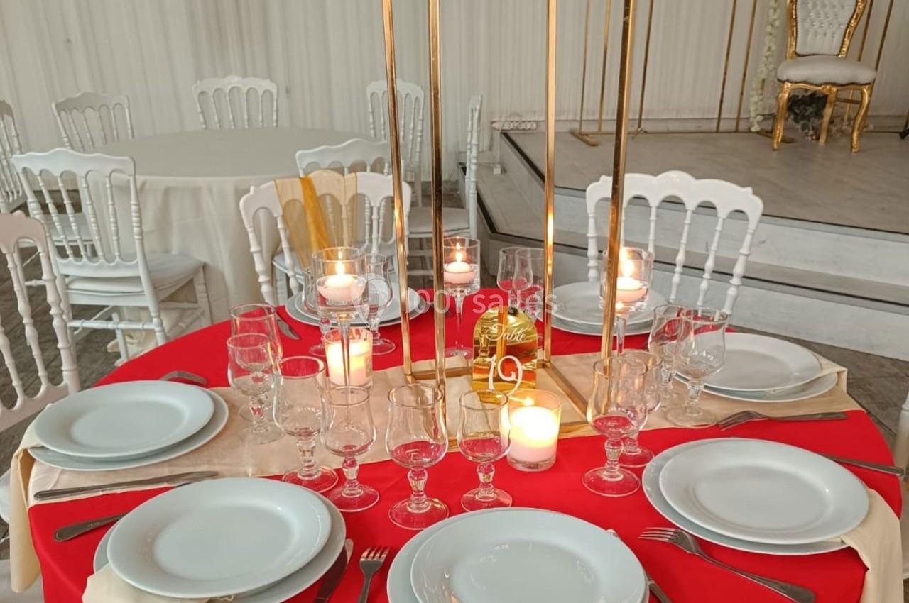Table dressée avec une nappe rouge, vaisselle blanche, verres, bougies allumées et décoration dorée dans une salle.