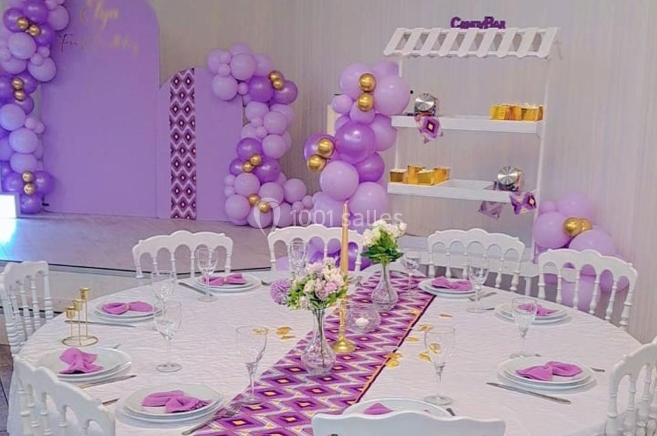 Table décorée avec nappe blanche, serviettes violettes, centre de table floral, et arrière-plan avec ballons violets et…
