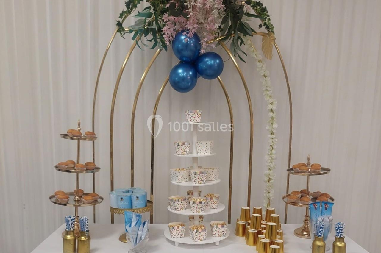 Table décorée pour une réception avec pâtisseries, gobelets dorés, ballons bleus et un présentoir central.