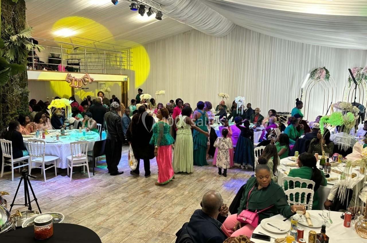 Des invités vêtus de tenues colorées participent à une réception dans une salle décorée avec des tables et des fleurs.
