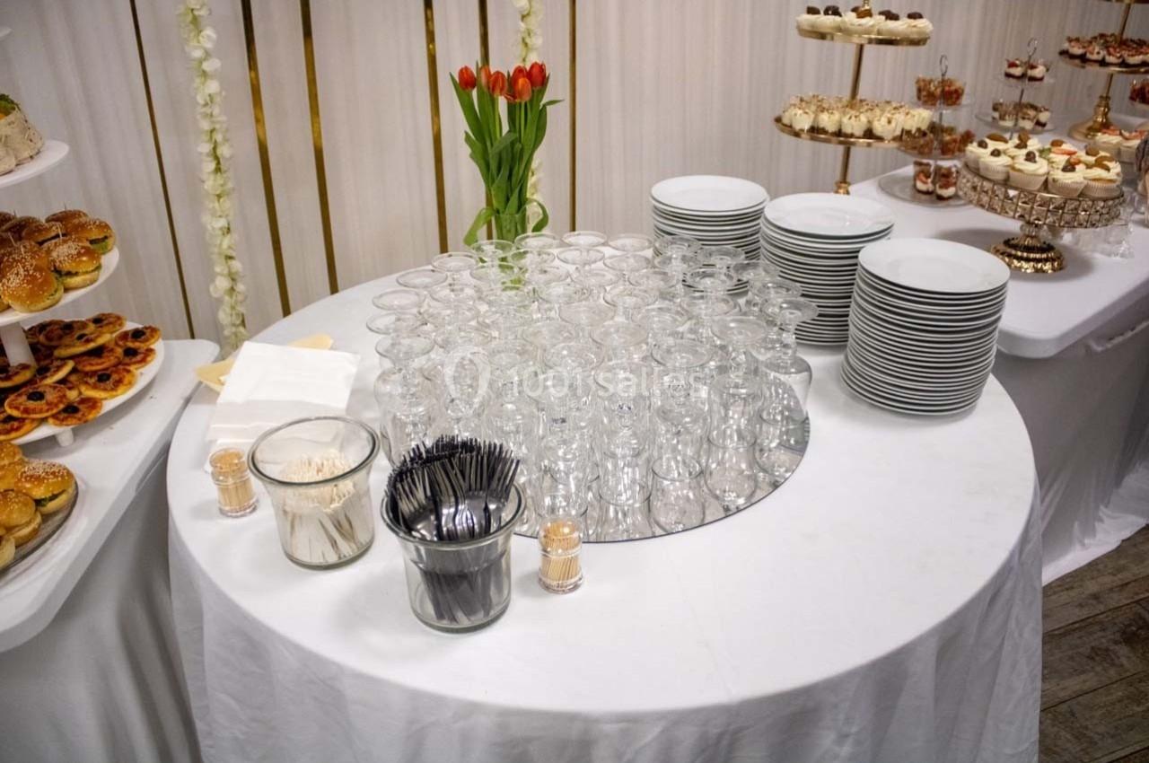 Table ronde avec verres vides, assiettes empilées, couverts, serviettes et desserts en arrière-plan.