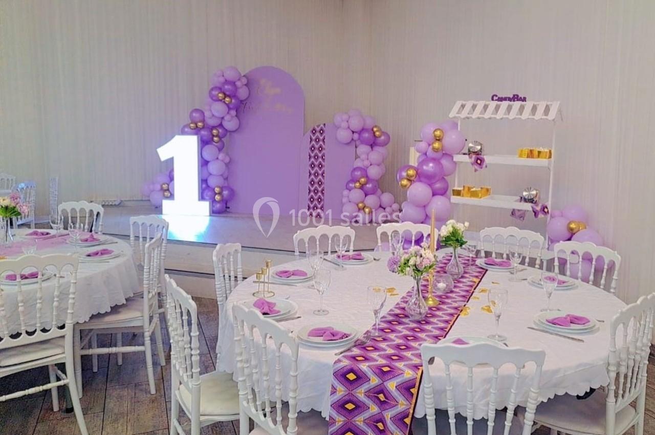 Salle décorée pour une fête avec tables dressées, ballons violets et dorés, et un grand chiffre ’1’ lumineux.