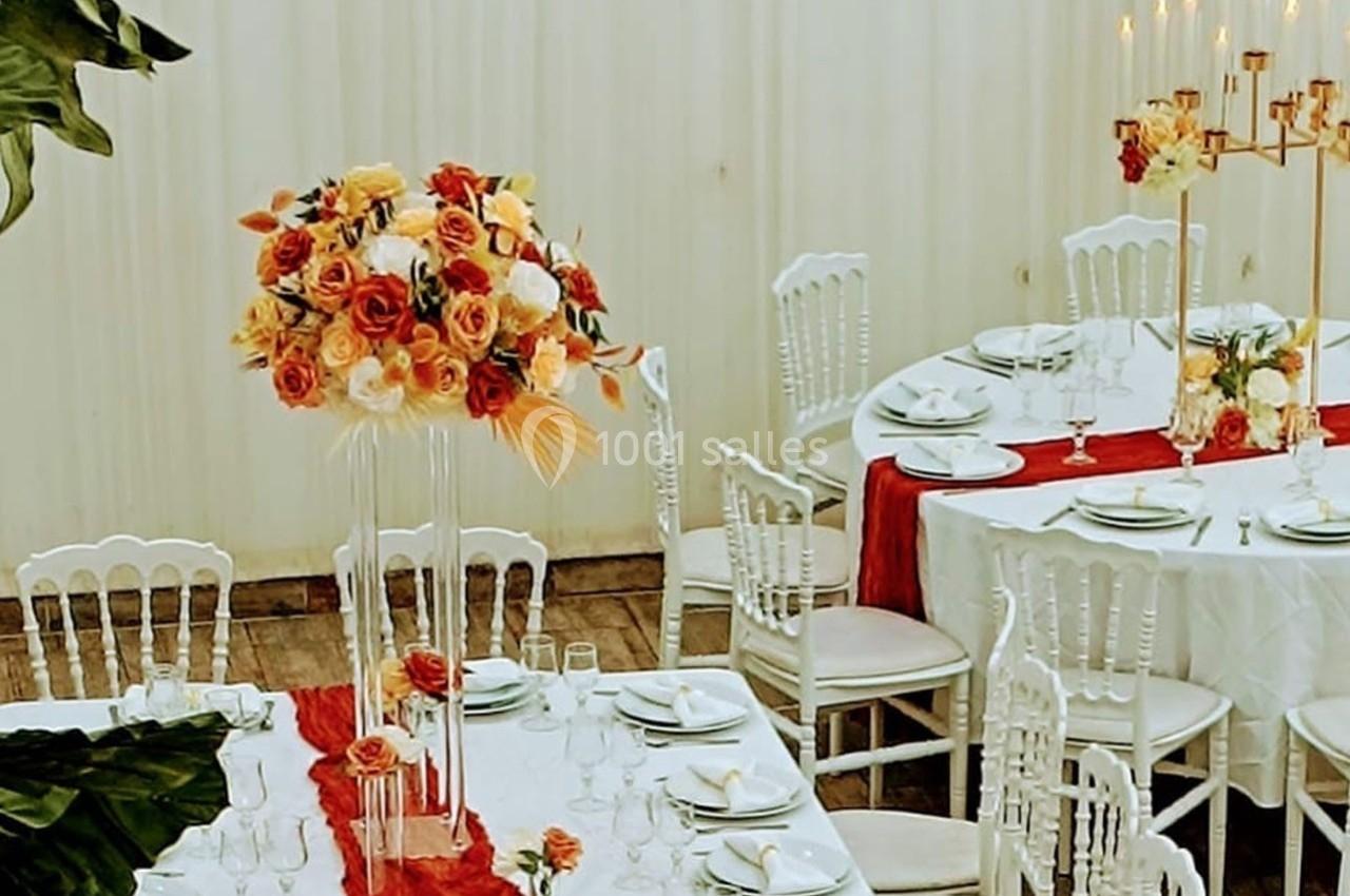 Salle décorée pour un événement avec tables dressées, nappes blanches, chemins de table rouges et bouquets de fleurs.