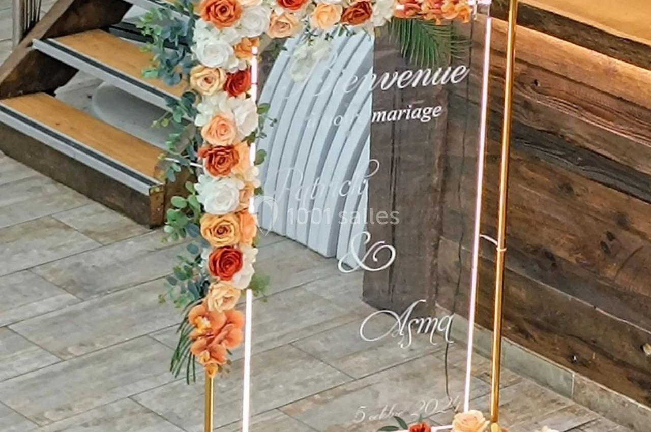 Panneau de bienvenue de mariage décoré de fleurs blanches et orange, posé sur un support doré.