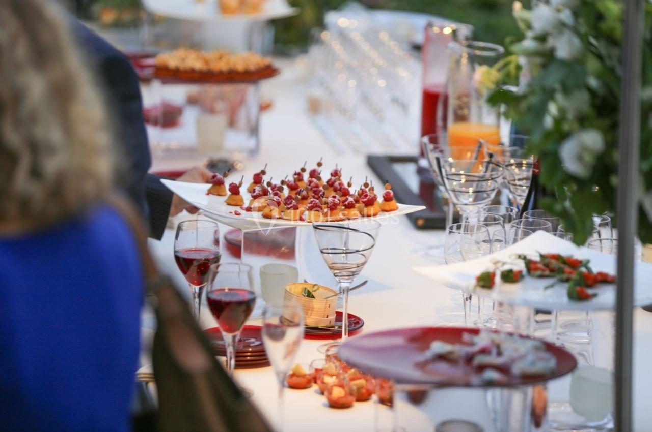Table dressée avec des amuse-bouches variés, verres de vin rouge et décorations florales en arrière-plan.