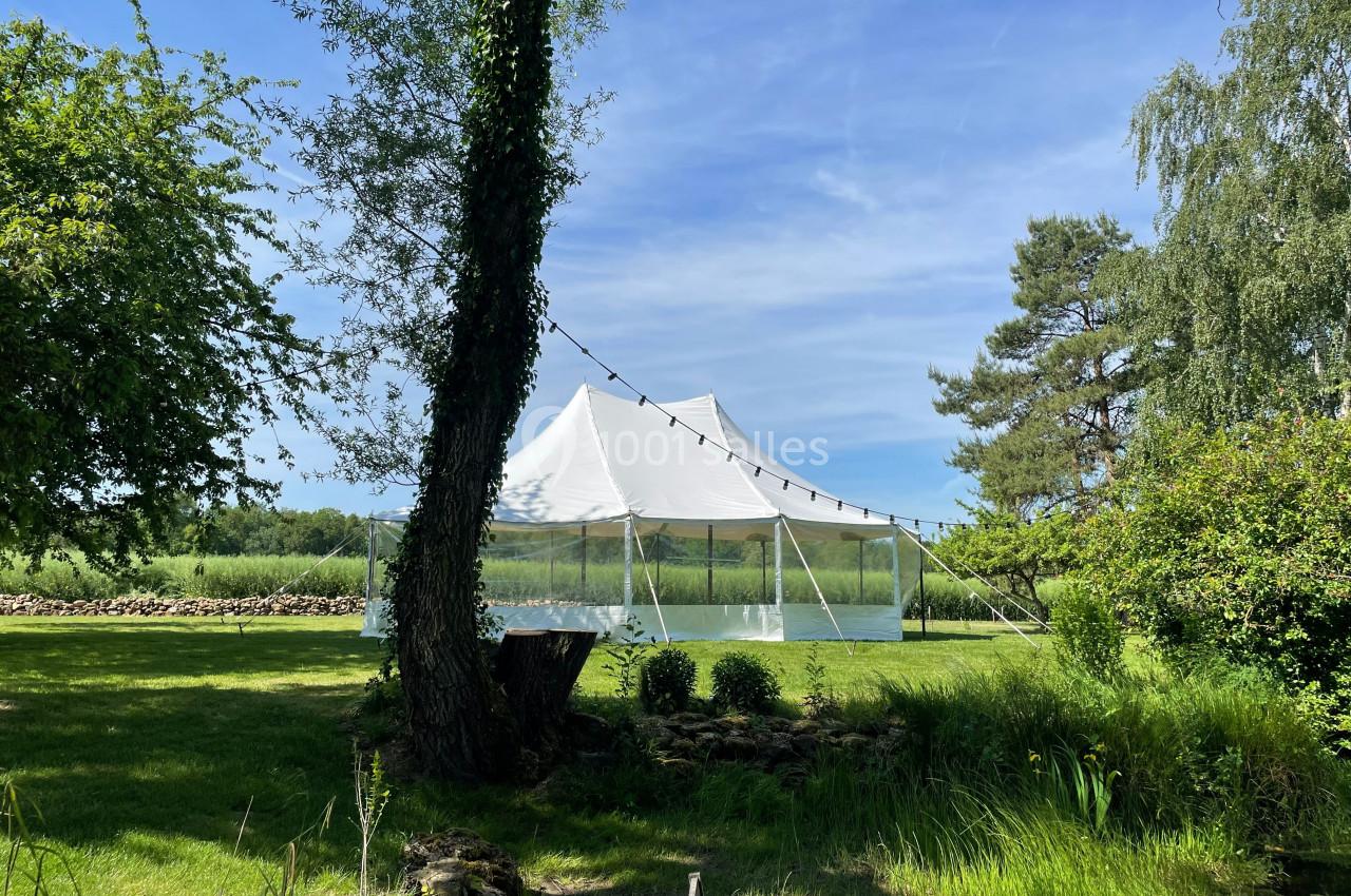 Tente blanche montée dans un jardin verdoyant, entourée d'arbres et éclairée par une guirlande lumineuse.