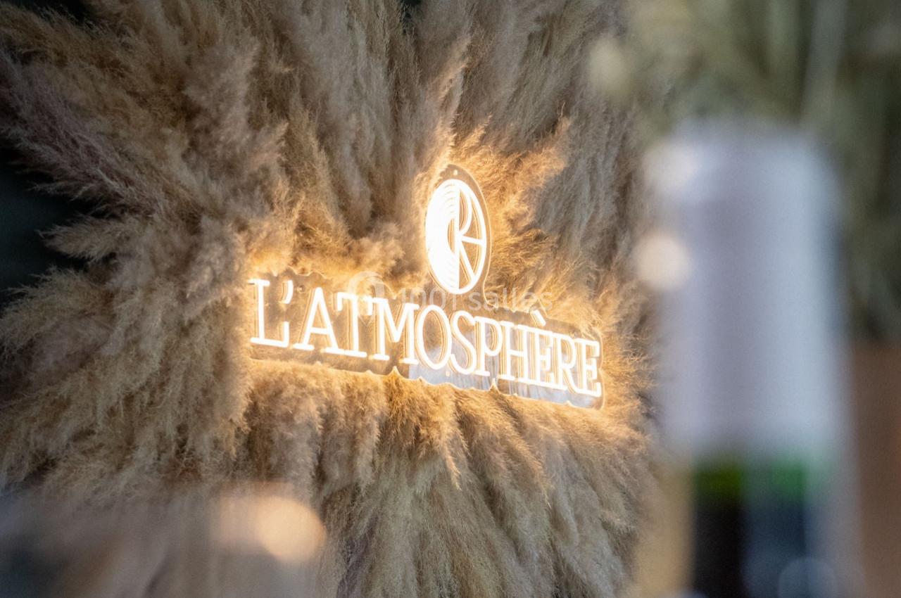 Enseigne lumineuse ’L'Atmosphère’ entourée de pampas beige sur un fond flou.