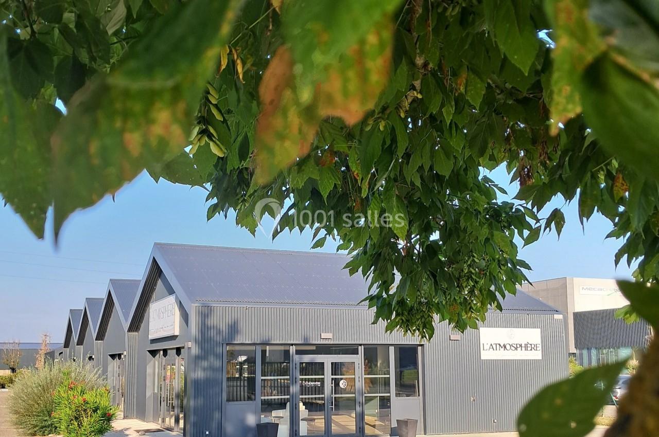 Bâtiment moderne en métal gris avec des grandes baies vitrées, partiellement caché par des feuilles d'arbre au premier plan.