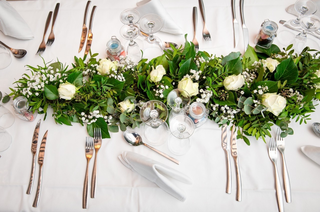 Centre de table avec des roses blanches, feuillage vert et vaisselle disposée sur une nappe blanche.