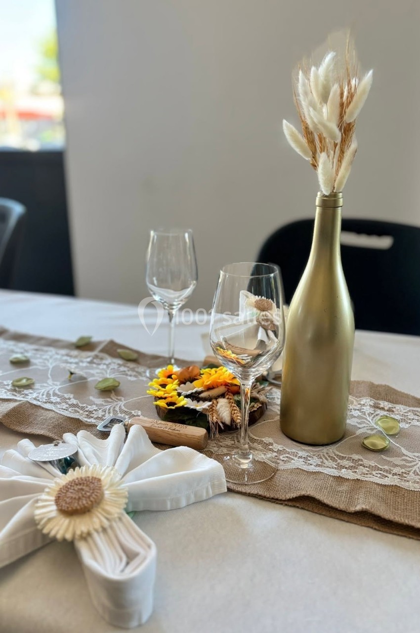 Décoration de table avec nappe blanche, chemin de table en jute et dentelle, bouteille dorée et fleurs séchées.