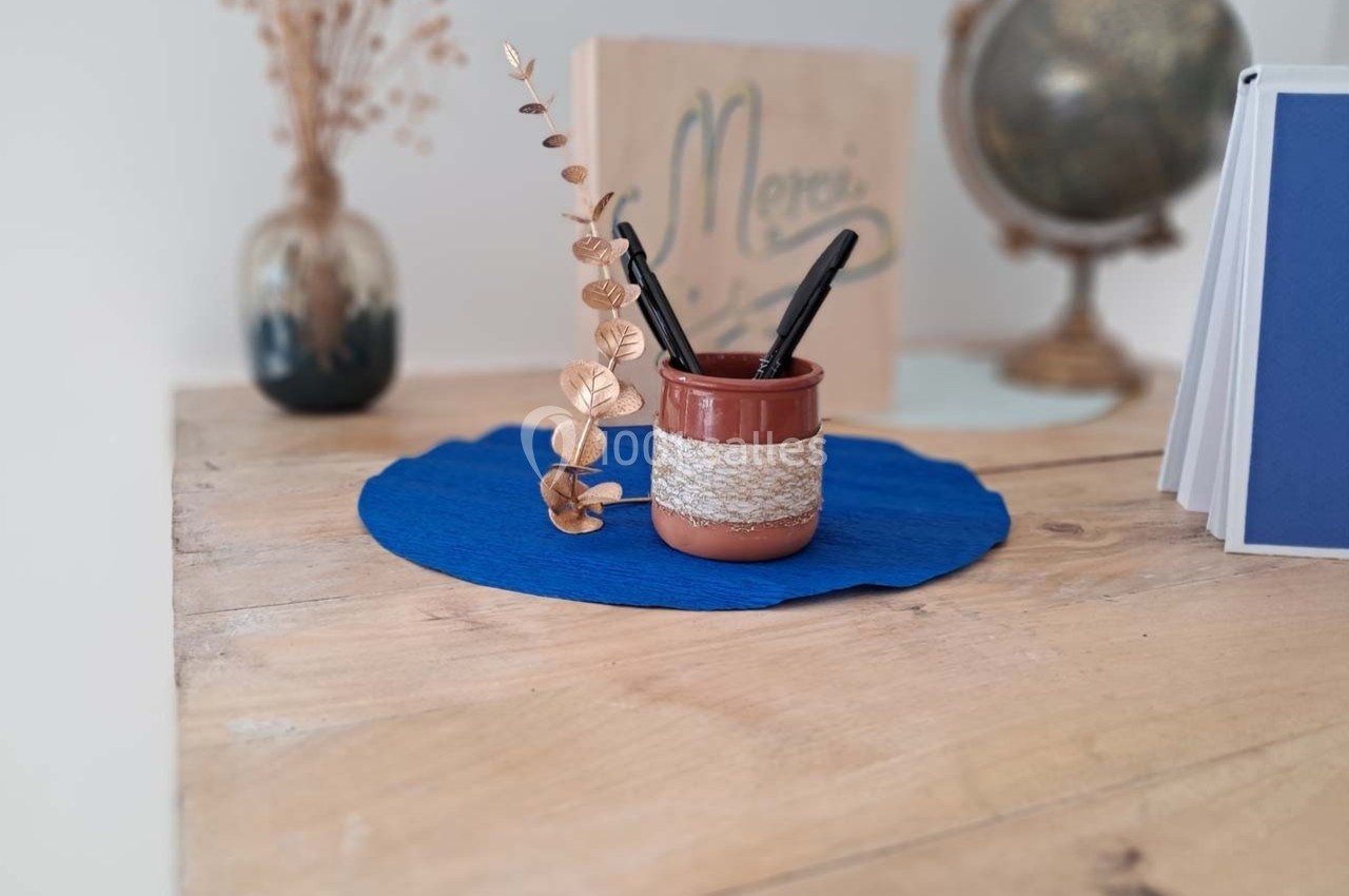 Pot en céramique contenant des stylos et une branche décorative, posé sur un napperon bleu sur une table en bois.