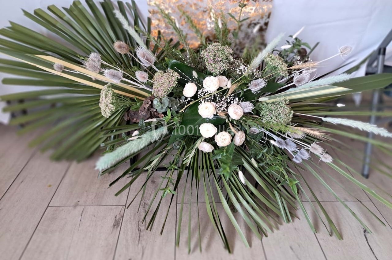 Composition florale avec des feuilles de palmier, des roses blanches et des fleurs séchées, posée sur un sol en bois clair.