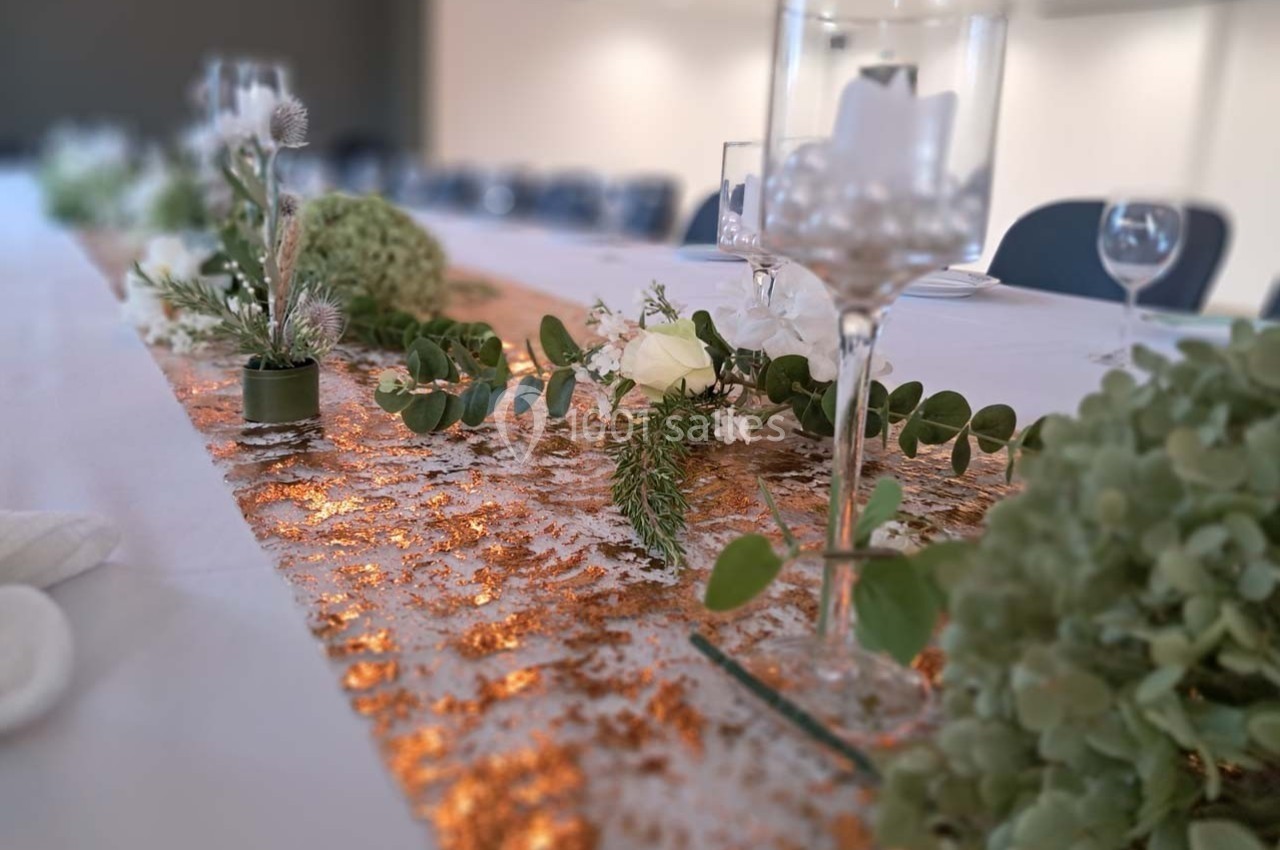 Décoration de table avec chemin doré, feuillage vert, fleurs blanches et verres élégamment disposés.