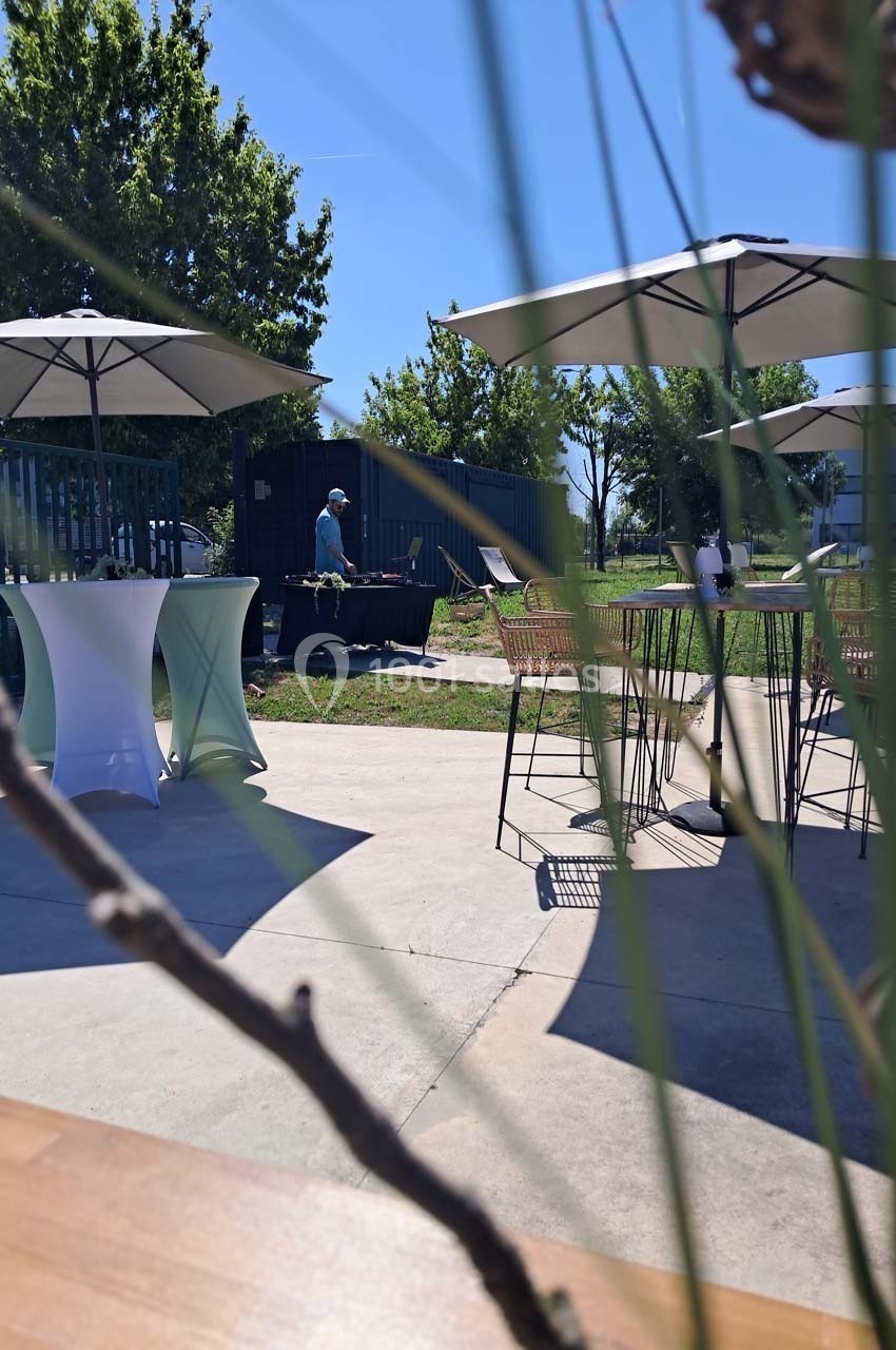 Espace extérieur avec tables hautes, parasols et un homme près d'une table, entouré de verdure sous un ciel dégagé.