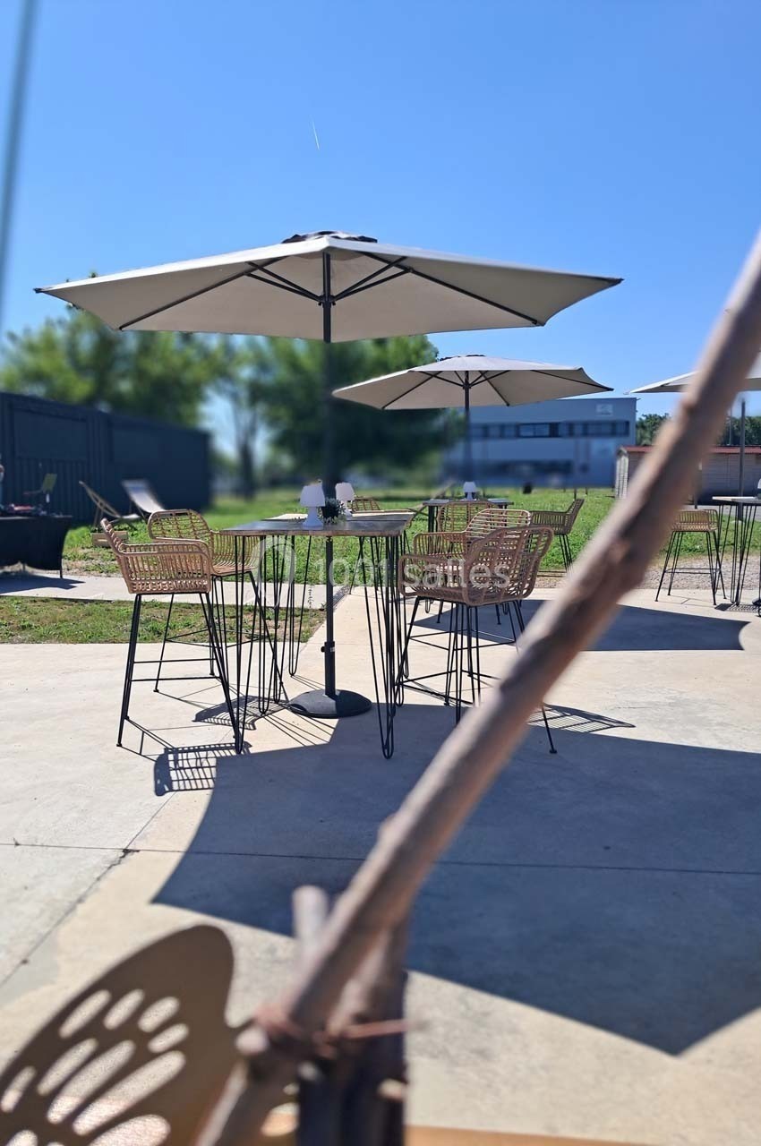 Terrasse ensoleillée avec tables, chaises en rotin et parasols, entourée d'herbe et de bâtiments en arrière-plan.