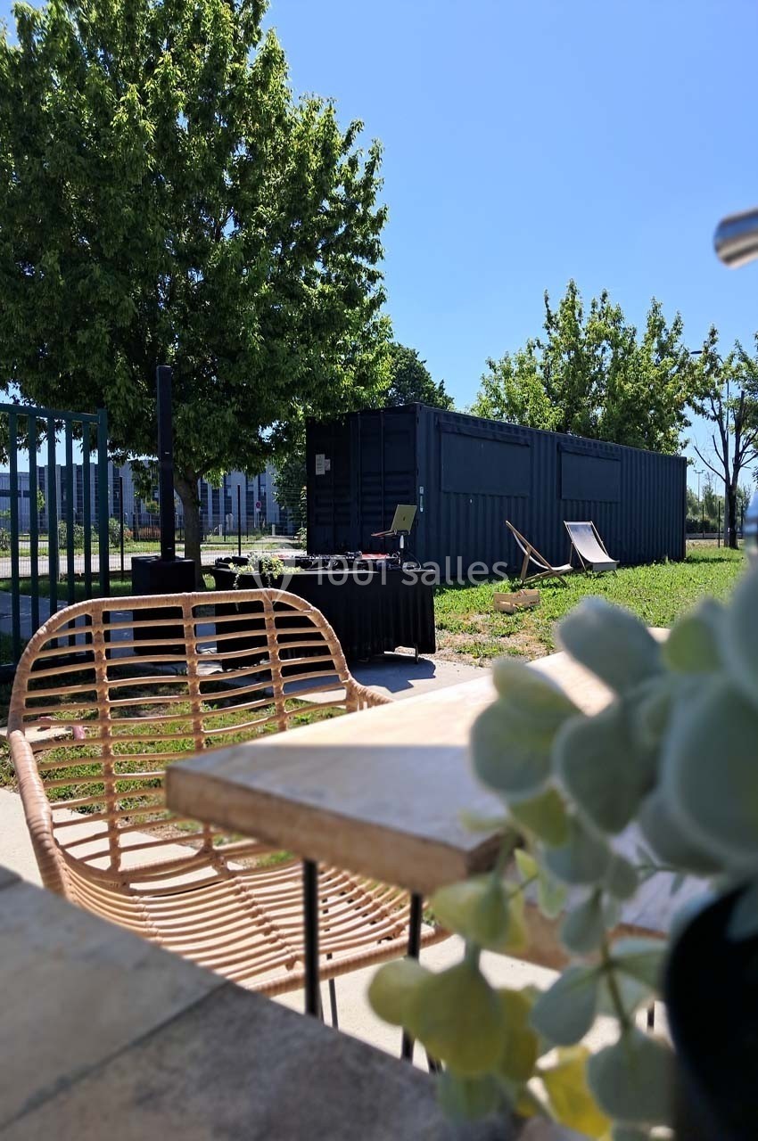 Table en extérieur avec chaise en rotin, plantes au premier plan, conteneur et arbres en arrière-plan sous un ciel bleu.