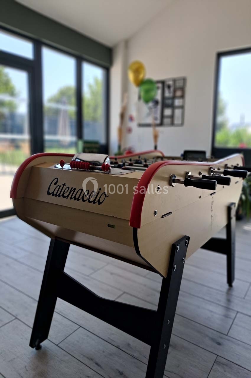 Table de baby-foot en bois clair avec des poignées rouges, située dans une pièce lumineuse avec de grandes fenêtres.