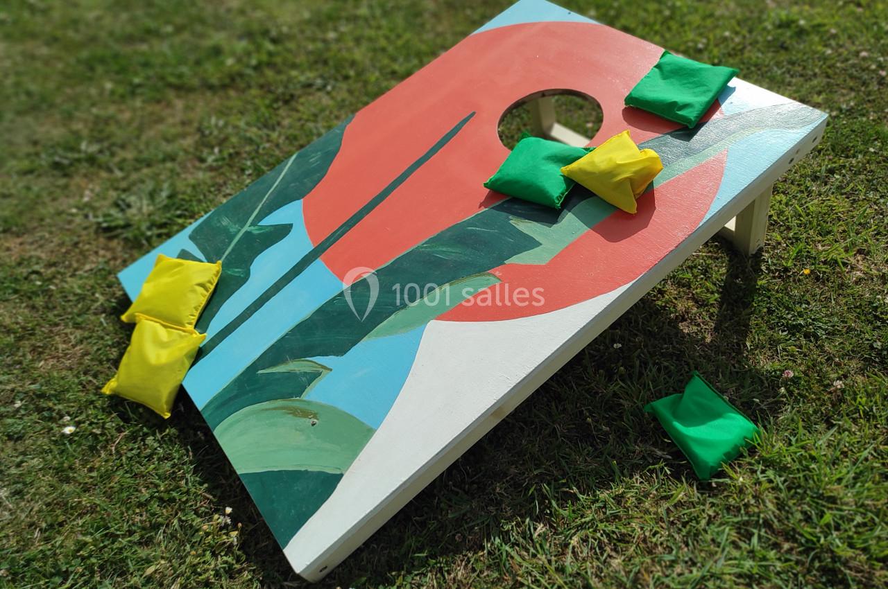 Planche de jeu de lancer de sacs colorée posée sur une pelouse, avec des sacs verts et jaunes dispersés autour.