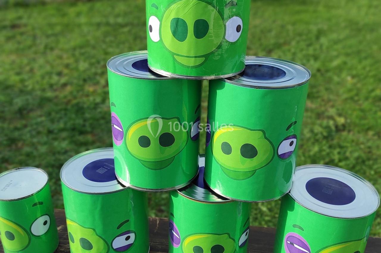 Six boîtes de conserve décorées de visages de cochons verts empilées en pyramide sur une table en extérieur.