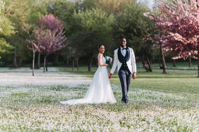 Un couple de mariés pose près d'une voiture ancienne blanche devant un bâtiment historique et des arbres.