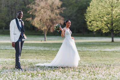 Un couple de mariés pose près d'une voiture ancienne blanche devant un bâtiment historique et des arbres.