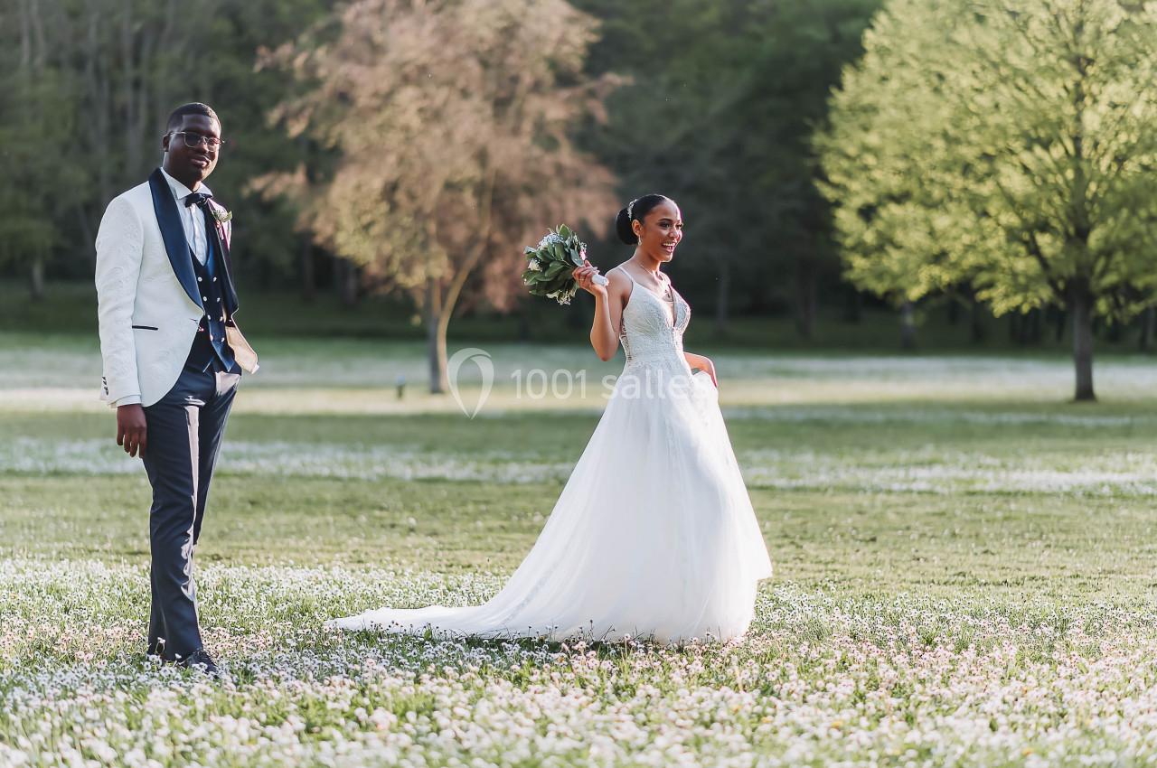 Un couple en tenue de mariage pose dans un champ fleuri avec des arbres en arrière-plan.