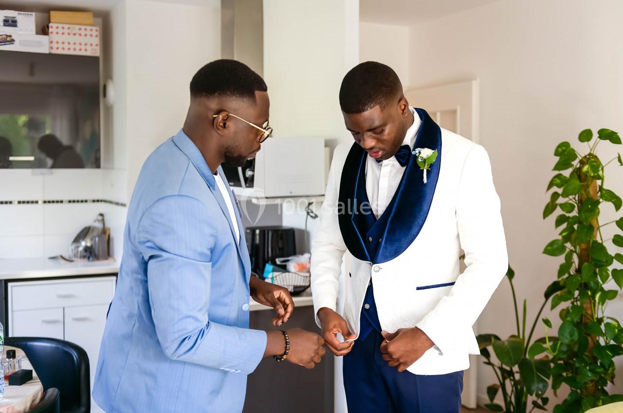 Deux hommes dans une cuisine, l'un en costume bleu clair aide l'autre à ajuster son costume blanc avec col bleu.