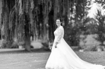 Un couple de mariés pose près d'une voiture ancienne blanche devant un bâtiment historique et des arbres.