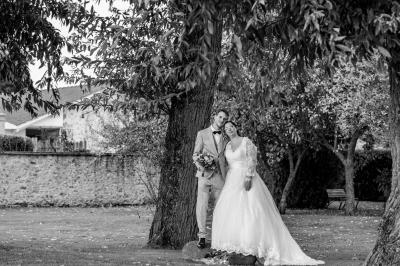 Un couple de mariés pose près d'une voiture ancienne blanche devant un bâtiment historique et des arbres.