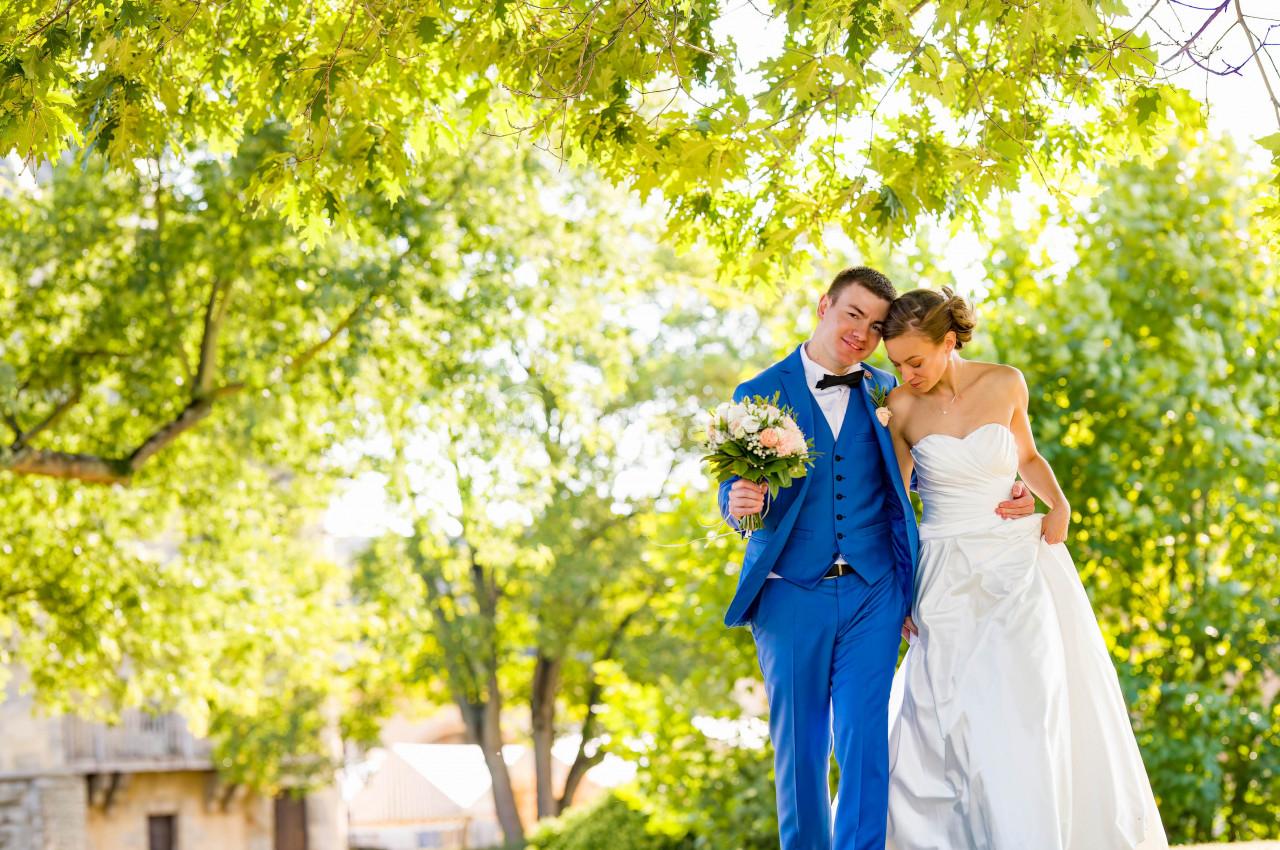 Un couple de mariés souriant, sous des arbres verdoyants, la mariée tenant un bouquet de fleurs.