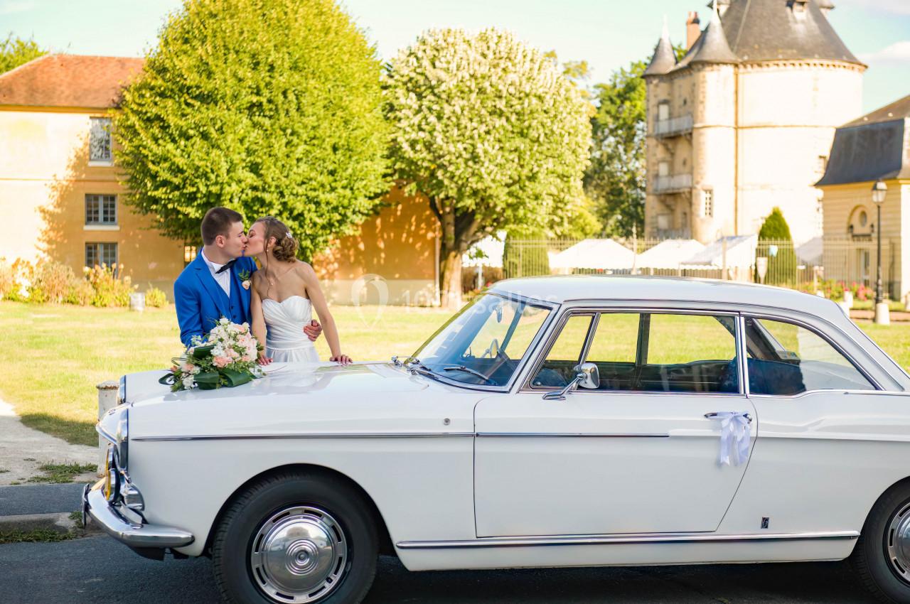 Un couple de mariés pose près d'une voiture ancienne blanche devant un bâtiment historique et des arbres.