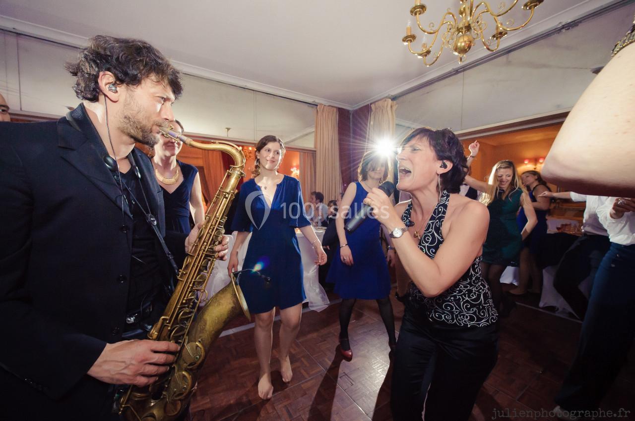 Une femme chante face à un saxophoniste lors d'une fête dans une salle éclairée, entourés de personnes dansantes.