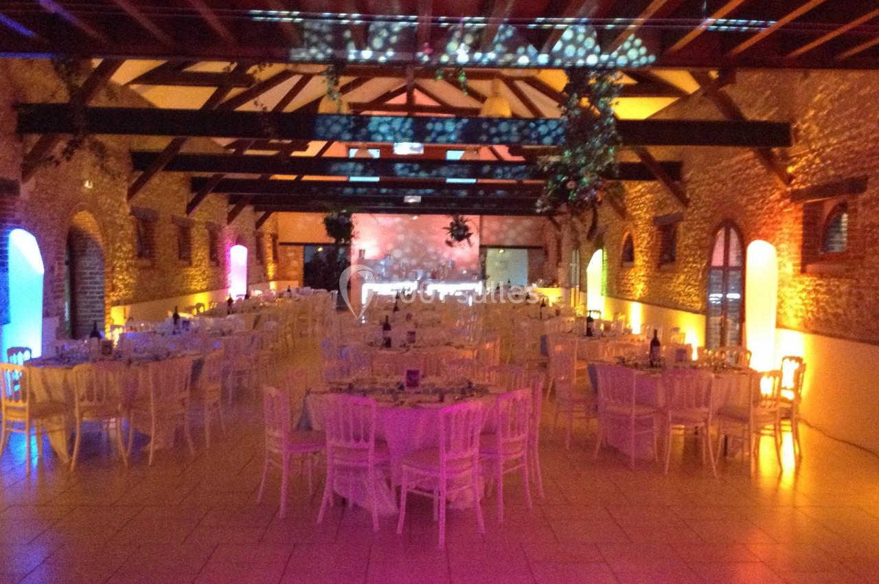 Salle de réception en pierre avec poutres apparentes, tables rondes dressées et éclairage coloré.