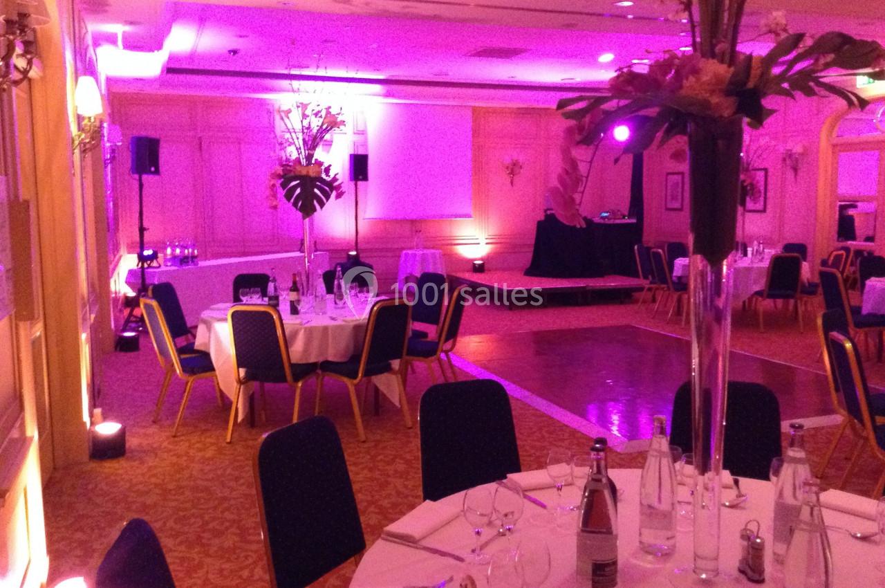 Salle de réception avec tables rondes dressées, éclairage tamisé rose et espace pour une scène.