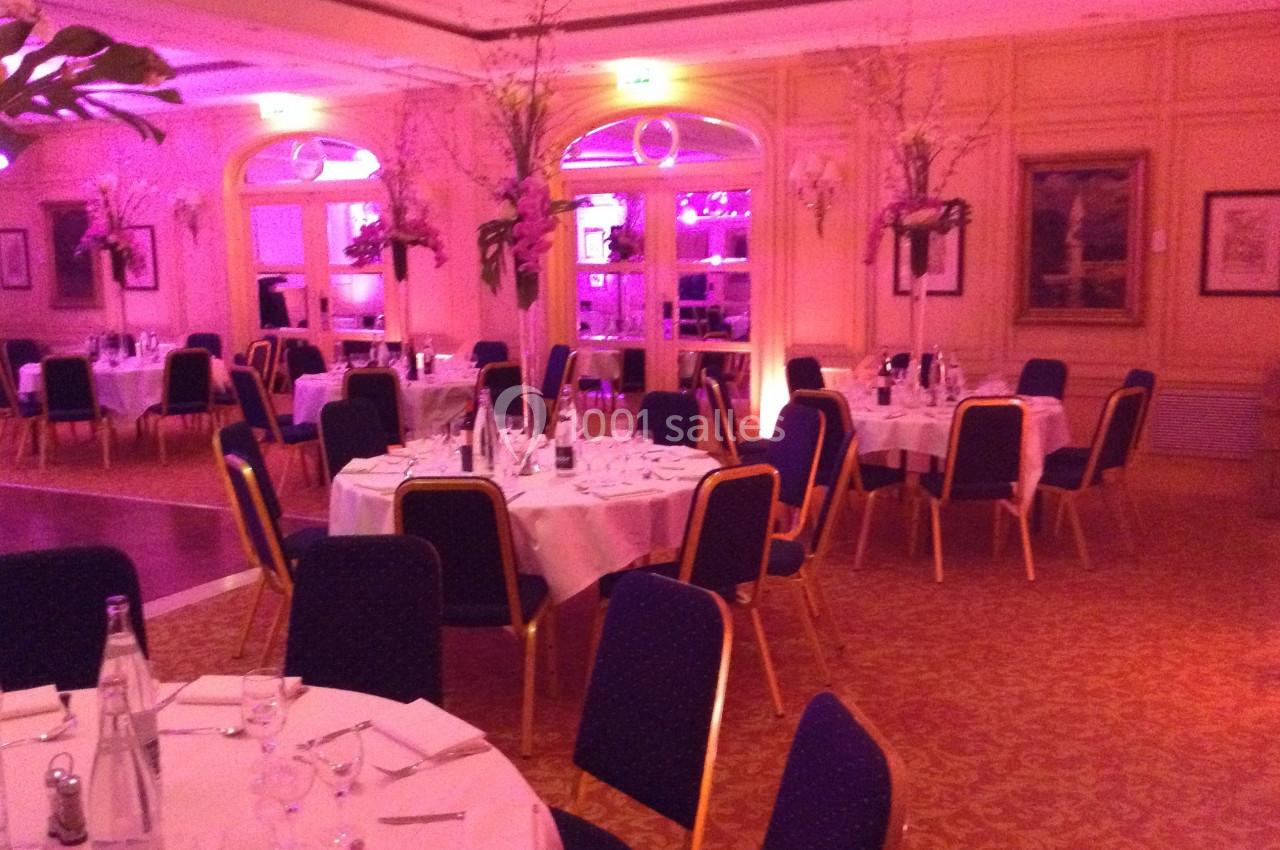 Salle de réception élégante avec tables rondes dressées, chaises bleues et éclairage tamisé rose.