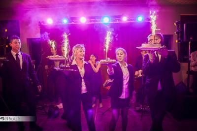 Des serveurs en tenue formelle apportent des plateaux avec des desserts ornés de cierges magiques dans une ambiance festive.