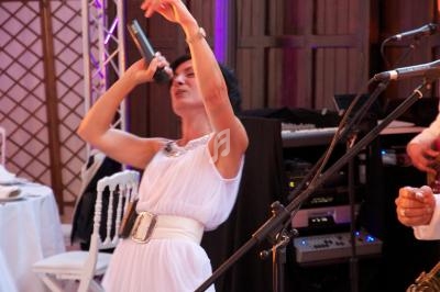 Femme en robe blanche chantant dans un micro lors d'un événement avec des équipements audio en arrière-plan.