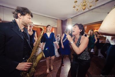 Un musicien joue du saxophone pendant qu'une femme chante, entourés de personnes dansant dans une salle éclairée.