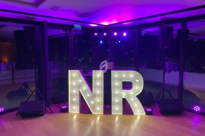 Lettres lumineuses ’NR’ posées sur un sol en bois, entourées de projecteurs et d'une baie vitrée en arrière-plan.