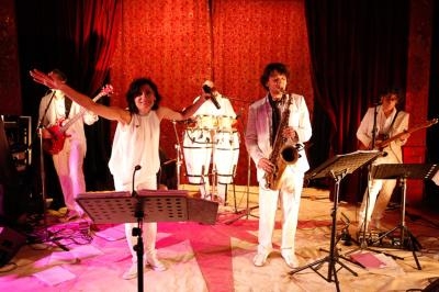 Groupe de musiciens en tenue blanche jouant sur scène avec éclairage rouge et instruments variés.