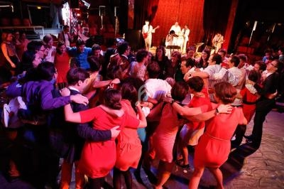 Groupe de personnes formant un cercle, bras dessus bras dessous, dans une salle éclairée avec des musiciens en arrière-plan.